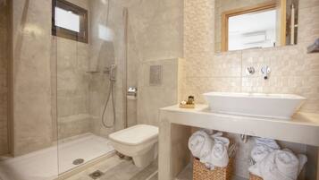 Un baño moderno con ducha de vidrio, inodoro blanco y lavabo con un recipiente amplio. Debajo del lavabo, hay dos cestas de mimbre con toallas.