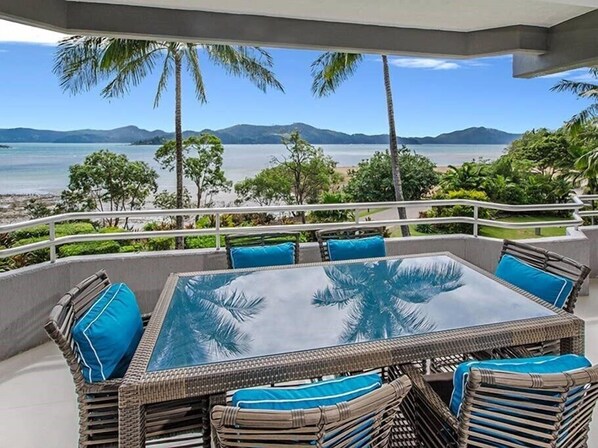 A glass-top table with wicker chairs and blue cushions, overlooking a beach and mountains. - Frangipani Lodge 006, Beachfront Apartment on Hamilton Island (Hamilton Island)
