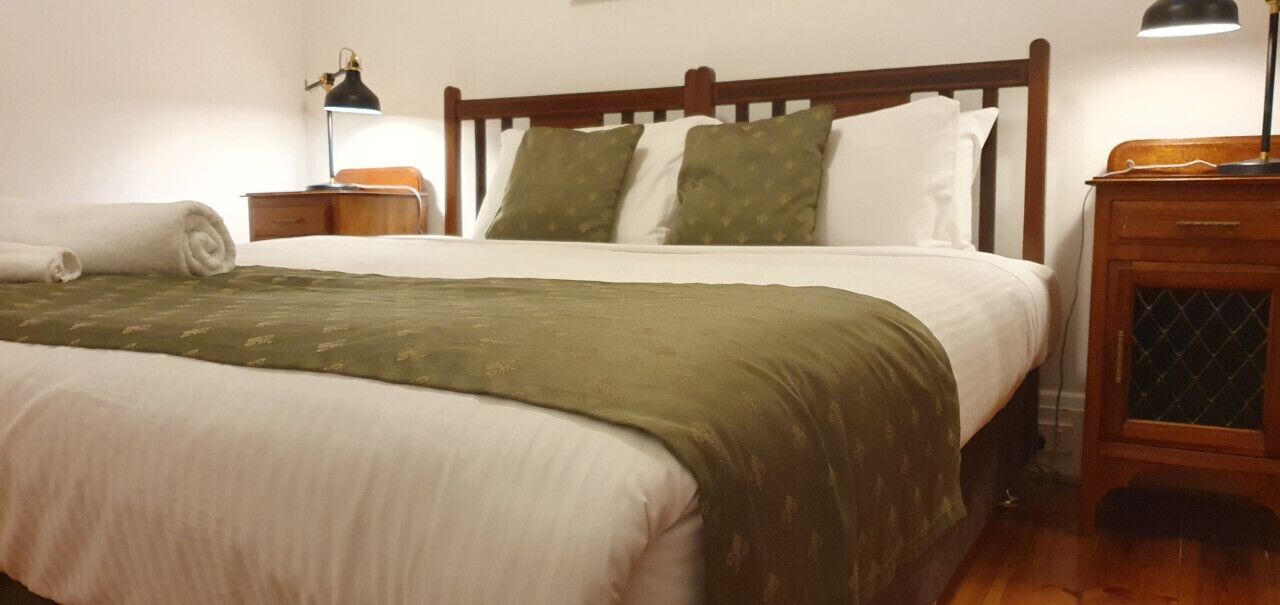 A neatly made bed with a green blanket, white pillows, and a wooden headboard. There are two bedside tables with lamps, one on each side of the bed.