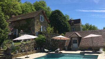 Une maison en pierre avec une piscine, entourée d’arbres et agrémentée de parasols d’extérieur.