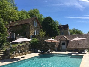 Une maison en pierre avec une piscine, entourée d’arbres et agrémentée de parasols d’extérieur.