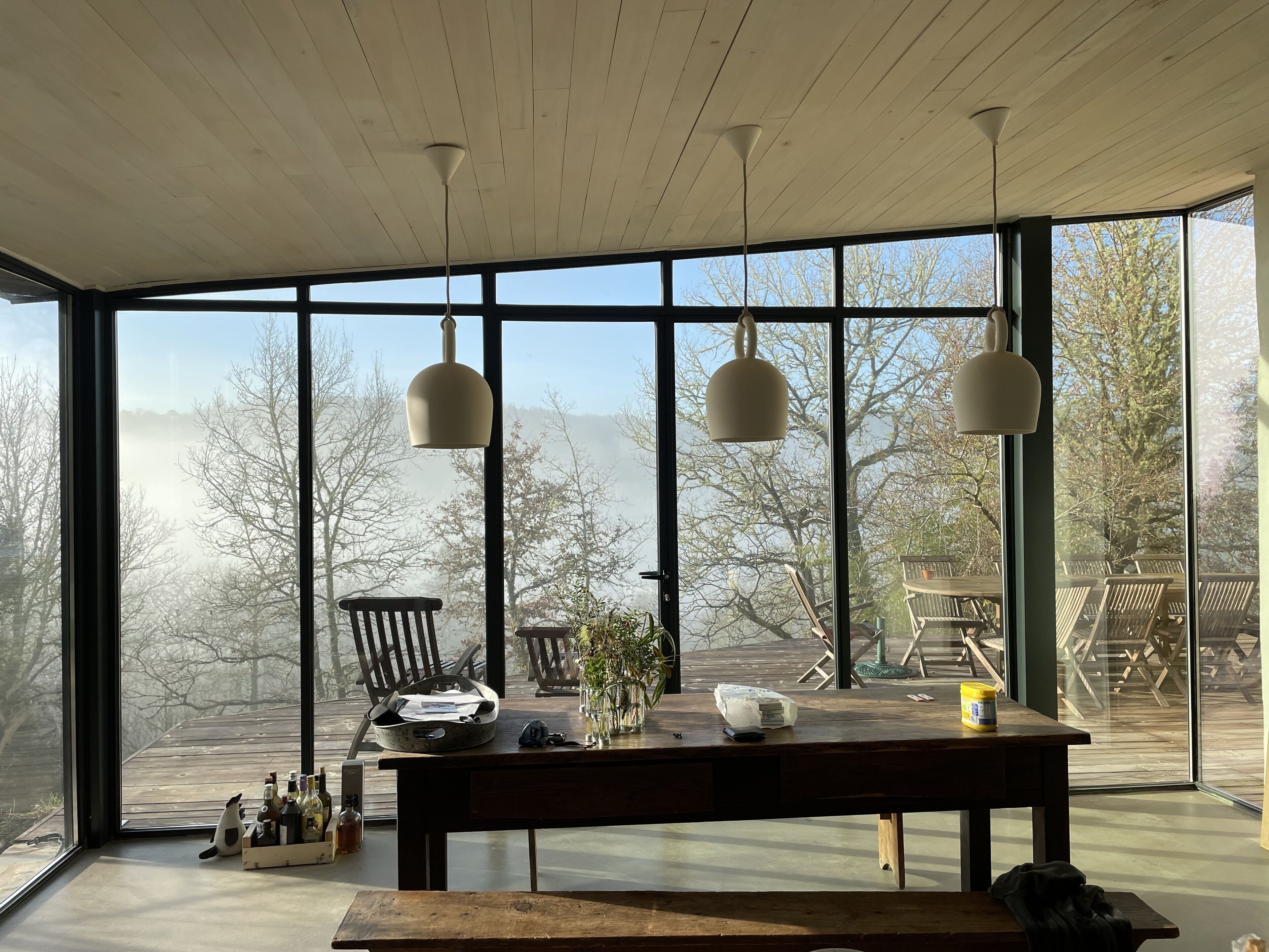 Une pièce spacieuse avec de grandes fenêtres, une table en bois sur laquelle sont disposés divers objets, et des lampes suspendues.