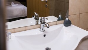 A hotel bathroom with a large mirror, a white sink, and a wooden door leading to a bedroom. - Hotel Garden (Novi Sad)