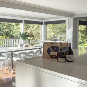 Una cocina moderna con una isla grande, encimeras blancas y vistas a zonas verdes. Hay dos botellas de vino y un frasco de mermelada sobre la isla.