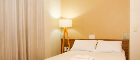 A hotel room with a bed, a desk, a chair, a lamp, and a curtain.