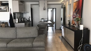 Een moderne woonkamer met een grijze bank, een eethoek met een tafel en stoelen, een keuken met een fornuis en kasten, en een groot abstract schilderij aan de muur.