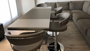 Een moderne eethoek met een rechthoekige tafel, twee stoelen met hoge rugleuningen en een grijze bank op de achtergrond.
