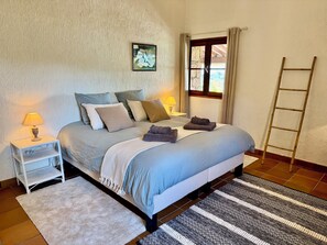 A neatly made bed with pillows and a blanket, a bedside table with a lamp, a window with curtains, and a wooden ladder against the wall.