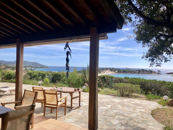 Un área de estar al aire libre con techo, suelo de piedra y mobiliario de madera, con vistas a un paisaje costero con colinas y una playa.