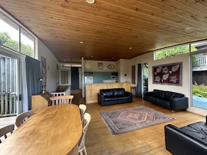 Amplia sala de estar con techo de madera, comedor con mesa y sillas de madera, sofá de cuero negro y cocina al fondo.