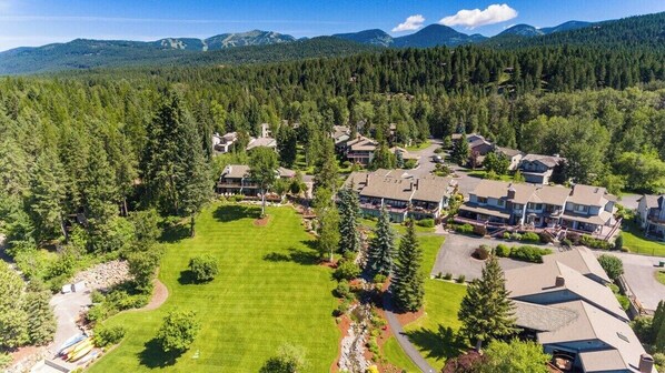 An aerial view of a residential area with houses, a central green space, and dense forested hills in the background.