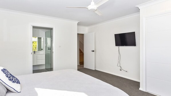 A modern bedroom with a bed, a ceiling fan, a TV mounted on the wall, and a door leading to a bathroom. - Heavenly Hideaway in Mollymook (Mollymook Beach)
