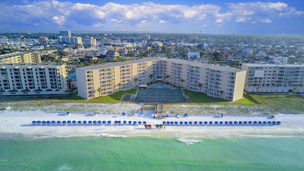 Un hotel frente al mar con varias edificaciones, una piscina y una playa con sombrillas.
