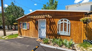 A small wooden building with a white door, a window with decorative ironwork, and a garden area with plants and a hanging flower pot. - Yellowstone's Treasure Cabin #1, few min drive to Yellowstone Park (Gardiner)