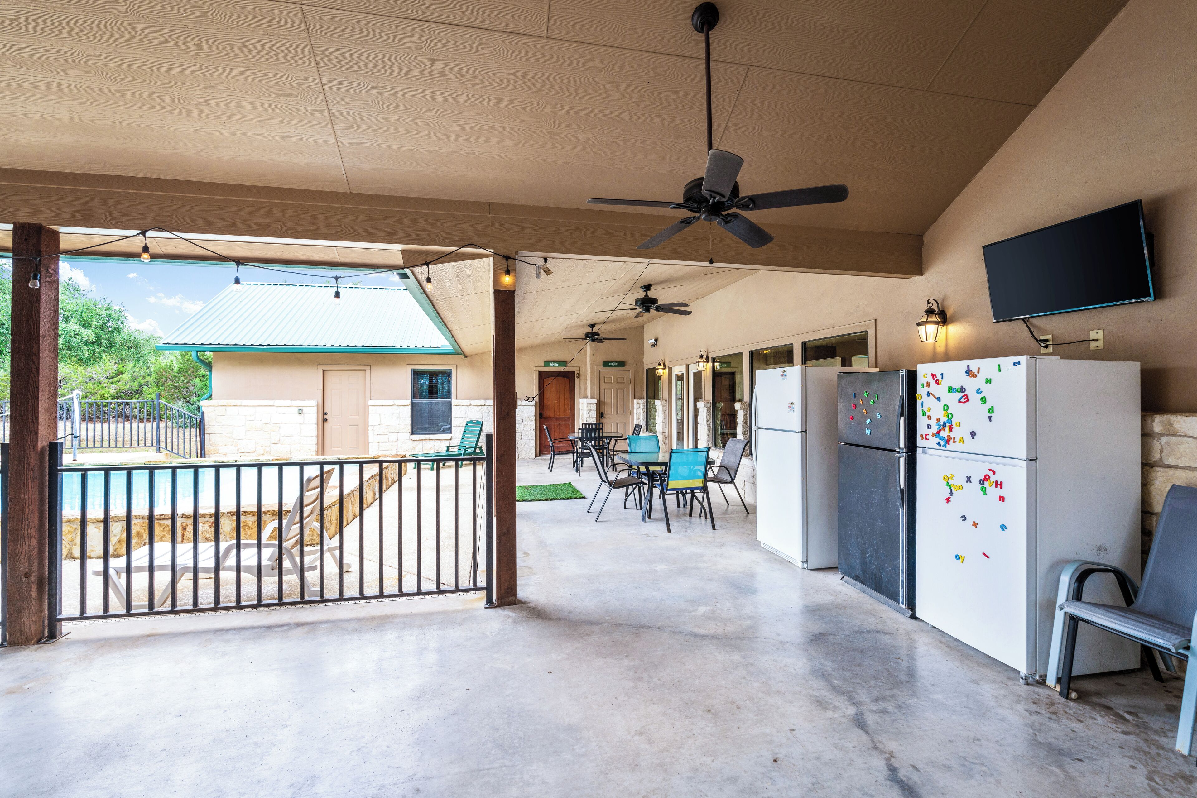 A covered outdoor area with a ceiling fan, a dining table with chairs, a refrigerator with magnets, and a TV mounted on the wall.