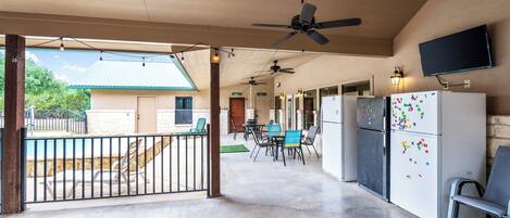 A covered outdoor area with a ceiling fan, a dining table with chairs, a refrigerator with magnets, and a TV mounted on the wall.
