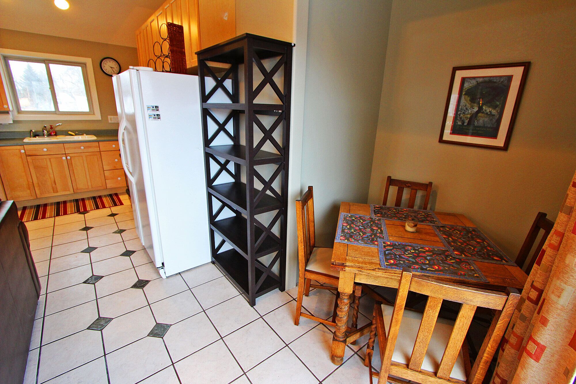 Una cocina con refrigerador, armarios de madera, piso de baldosas, una mesa de comedor con sillas y una repisa en la pared.
