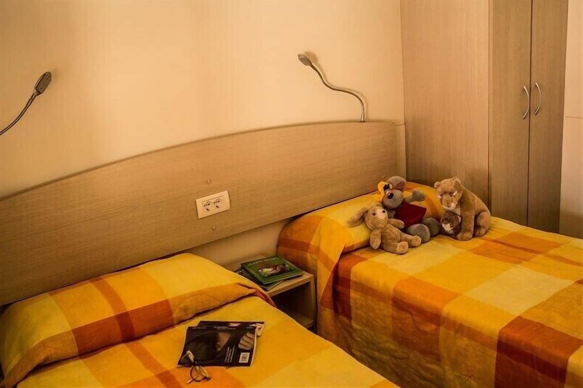 Ein Hotelzimmer mit zwei Betten, jeweils mit einer karierten Bettdecke, einem Nachttisch mit Buch und Gläsern sowie einem Kleiderschrank mit Spiegel.