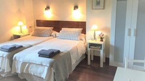 Habitación de hotel con dos camas, cada una con una almohada y una toalla doblada. Hay lámparas de noche y una mesita con una lámpara y una planta.