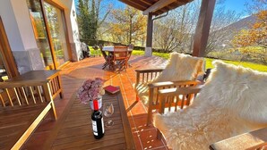 Un patio techado al aire libre con mobiliario de madera, una mesa con una botella y un vaso, y vistas a árboles y colinas.