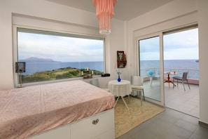 A bedroom with a large window overlooking the sea, a bed, a small table with a vase, and a chair.