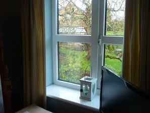 A window with a view of a garden, a white lantern, and a black object on the windowsill.