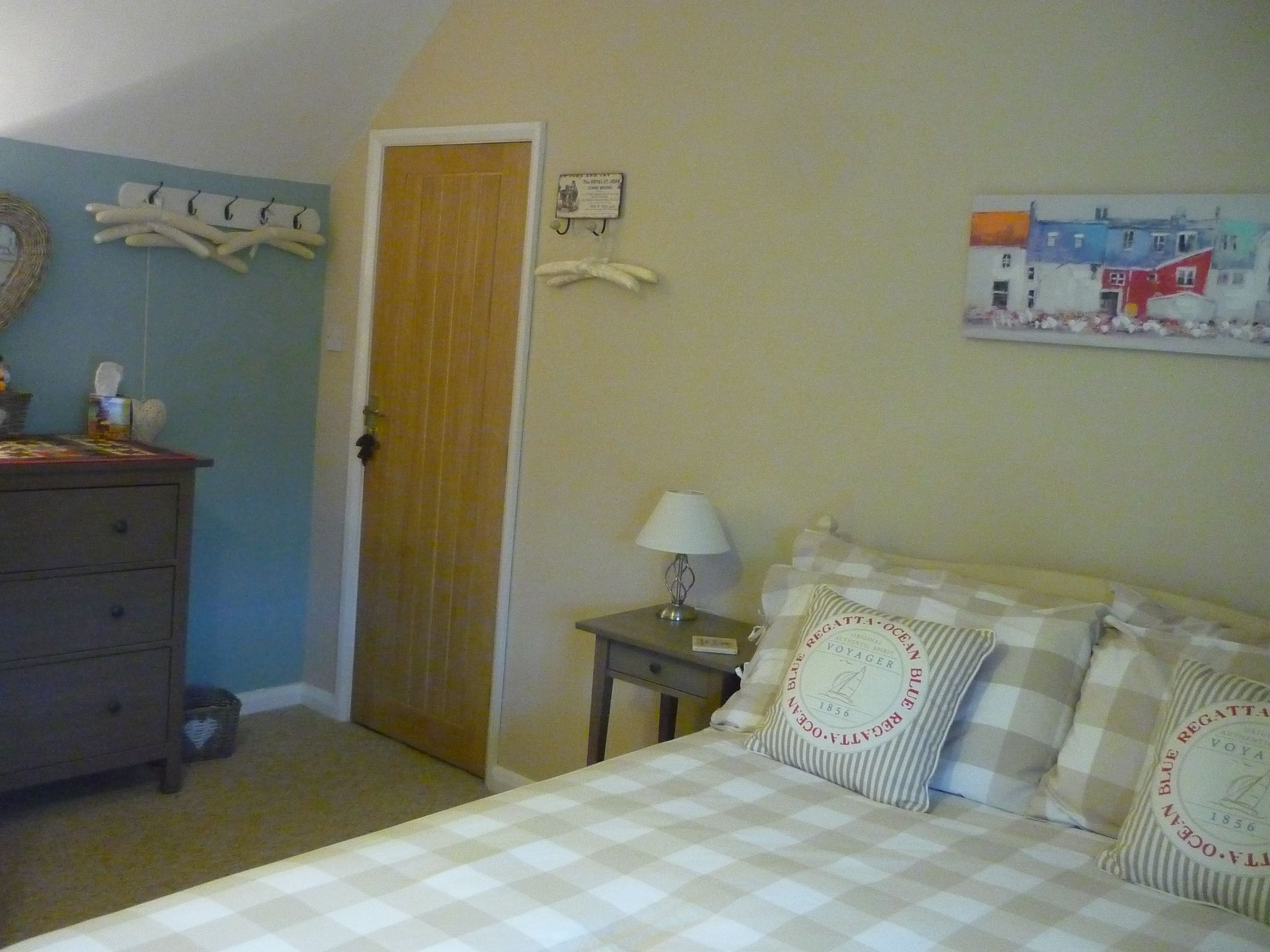 A bedroom with a checkered bedspread, a bedside table with a lamp, a dresser, a door, and a wall with a painting.