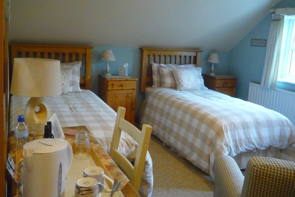 A bedroom with two beds, a wooden nightstand with a lamp, a small table with a kettle, and a chair.