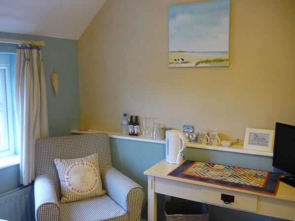 A cozy room with a checkered armchair, a small table with a kettle, glasses, and bottles, and a painting of a beach scene on the wall.