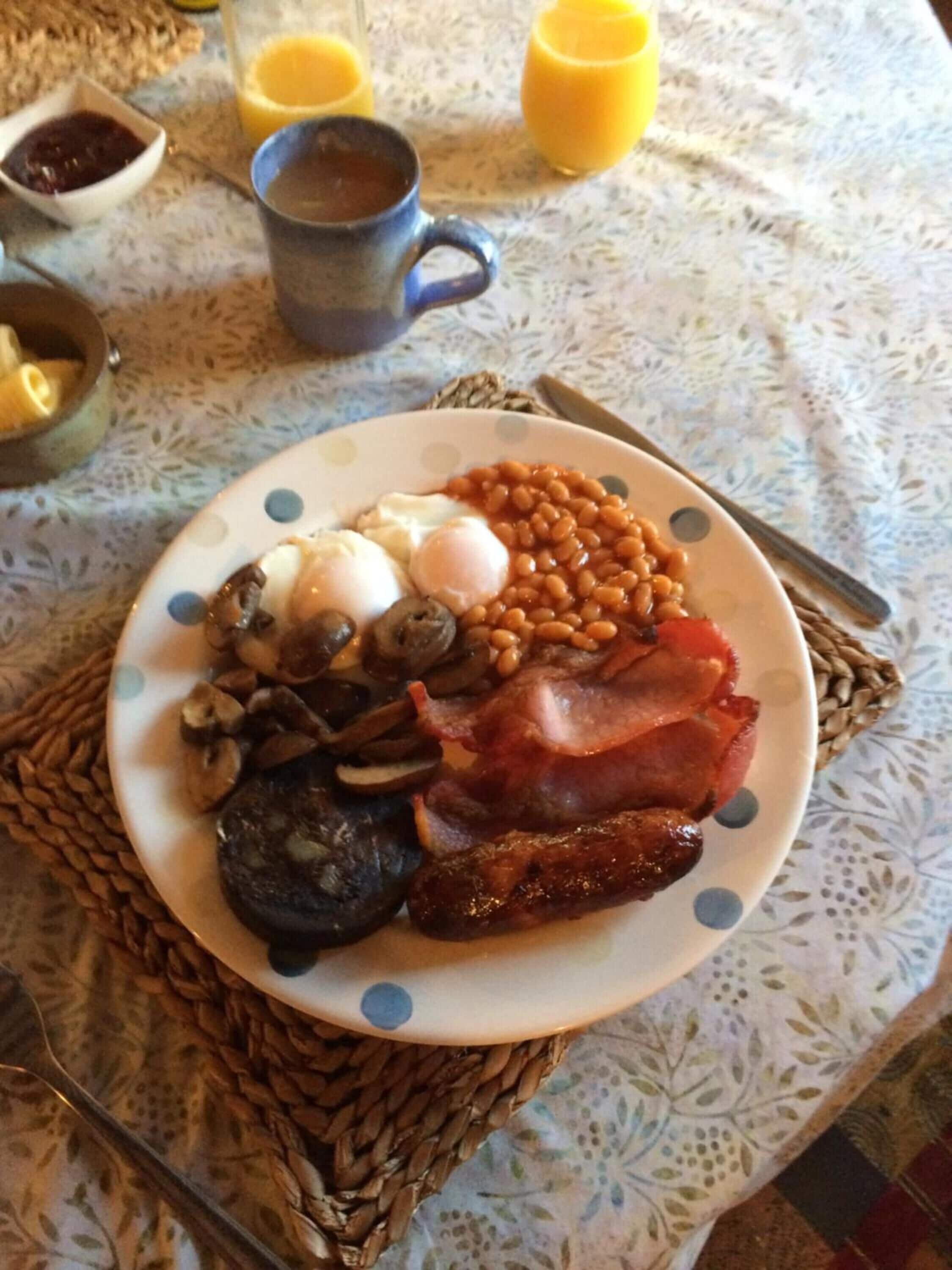Un plato de comida para el desayuno que incluye huevos, frijoles, champiñones, tocino y salchichas sobre una mesa con un mantel tejido.