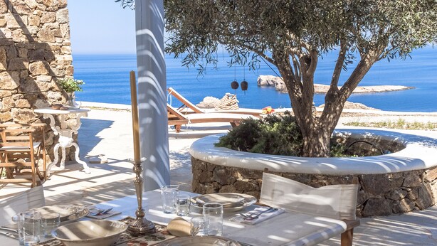 An outdoor dining area with stone walls, a table set with plates, glasses, and cutlery, and a view of the sea. - 200μ από τη παραλία  (MYKONOS)