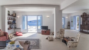 A spacious living room with a large sliding glass door leading to a balcony with a view of the sea. There is a television on a wooden stand, a red sofa, and a coffee table with a book and a bowl of fruit. - 200μ από τη παραλία  (MYKONOS)