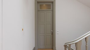 Een smalle gang met een gesloten deur aan het einde, een houten trap aan de rechterkant en een lichtschakelaar op de muur.