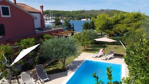 A pool area with lounge chairs, a red building, and a view of the sea and hills.