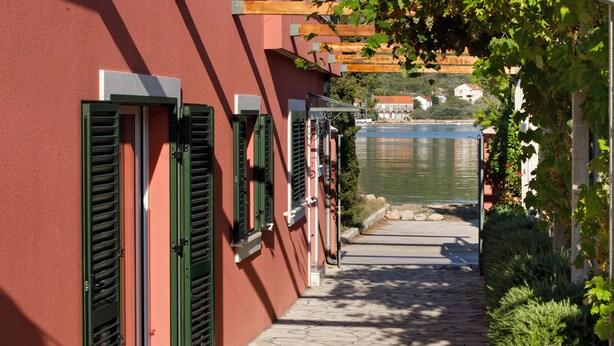 A narrow stone pathway leading to a building with green shutters and a view of a body of water.