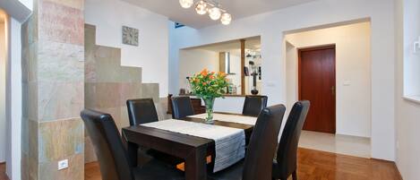 Un coin repas avec une table en bois, des chaises noires et un vase de fleurs. Un escalier se trouve à gauche et une porte à droite.