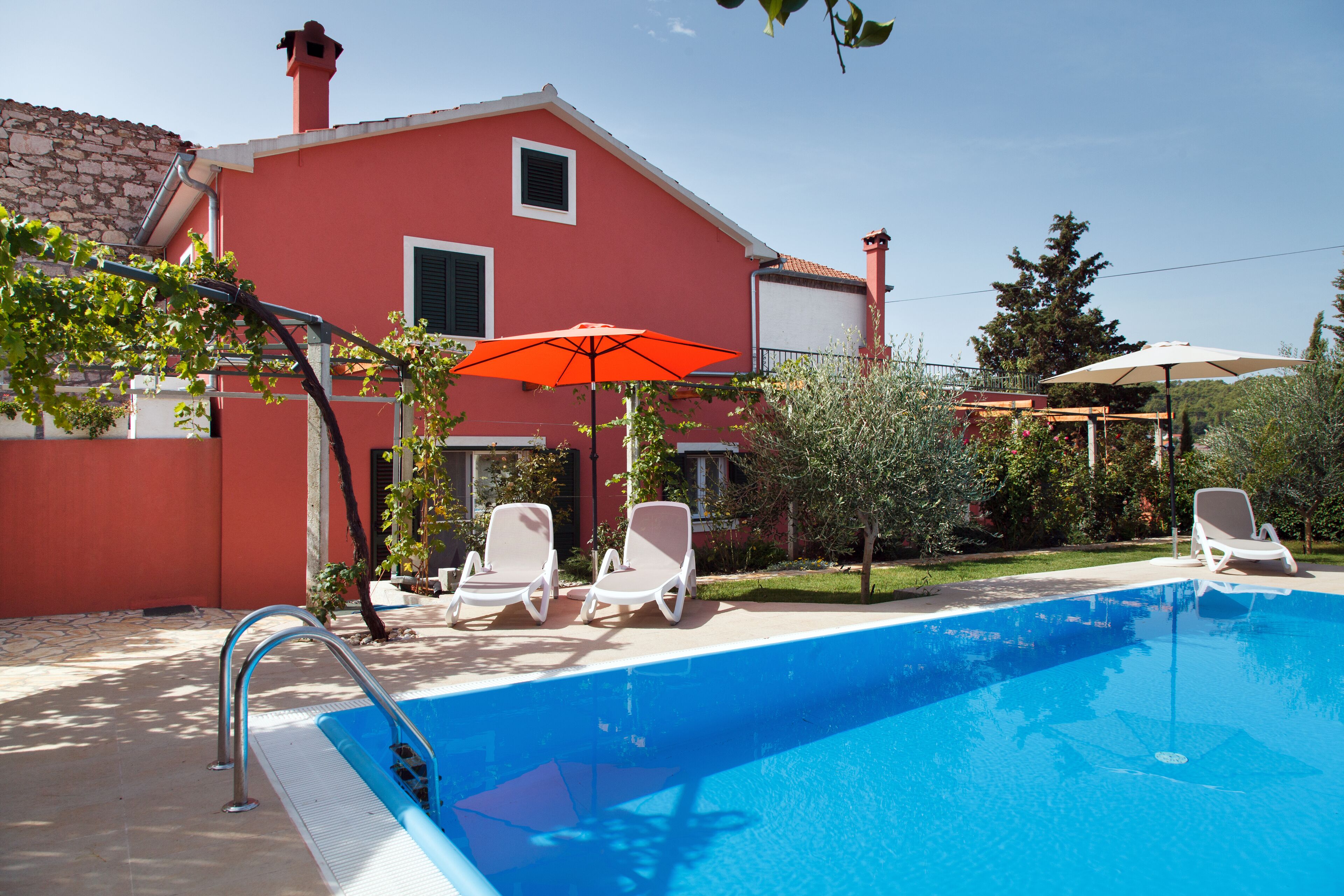 A swimming pool with two lounge chairs and an umbrella, surrounded by a red building and greenery.
