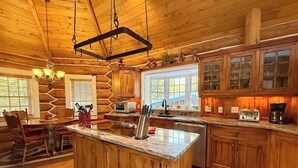 A log cabin interior with a central island, wooden cabinets, and a dining area with chairs. - Spectacular 4 Bedroom Log Home near Bootjack Lake (Whitefish)
