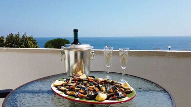 Une table sur laquelle on a disposé un plateau de fruits de mer, une carafe et deux coupes de champagne, le tout sur un balcon donnant sur la mer.