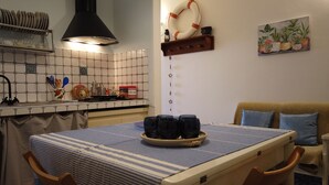 Une cuisine avec un mur recouvert de carreaux, une cuisinière et une table à manger recouverte d’une nappe bleue. Des chaises sont disposées autour de la table, et une étagère se trouve au-dessus de la cuisinière, où sont rangés des ustensiles de cuisine.