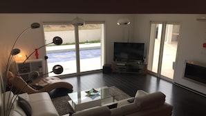 Un salon moderne avec un canapé blanc, une table basse en verre et un téléviseur à écran plat. Une cheminée se trouve à droite, et une porte-fenêtre coulissante donne accès à un espace extérieur.