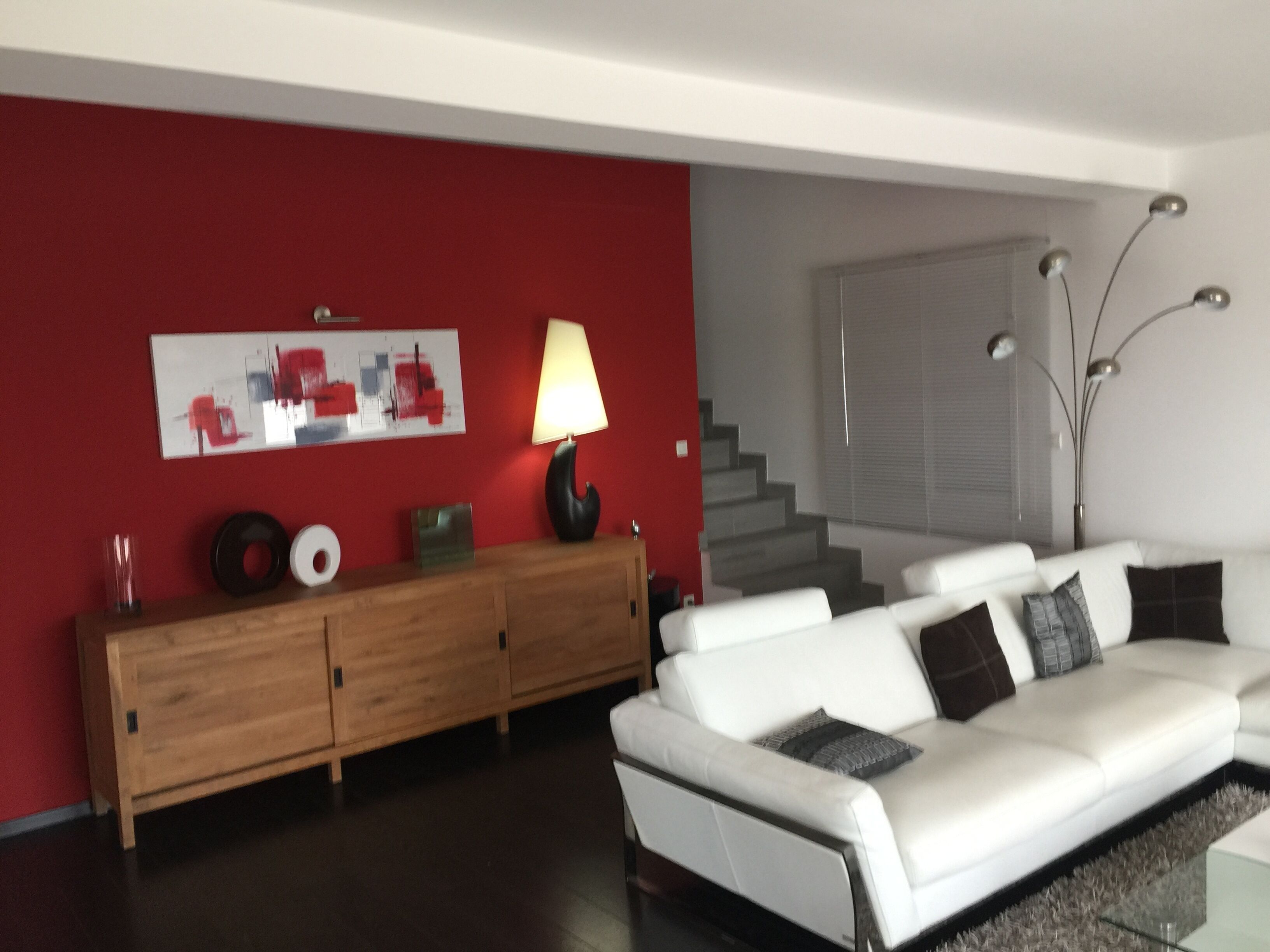 A modern living room with a white sofa, a wooden cabinet, a red accent wall, and a staircase in the background.