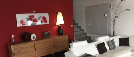 A modern living room with a white sofa, a wooden cabinet, a red accent wall, and a staircase in the background.
