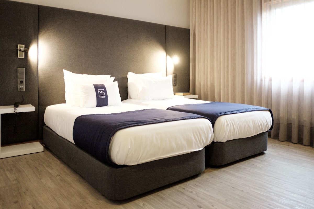 Un lit double avec une tête de lit sombre et du linge de lit blanc, une table de chevet avec une lampe, et une fenêtre avec des rideaux légers.