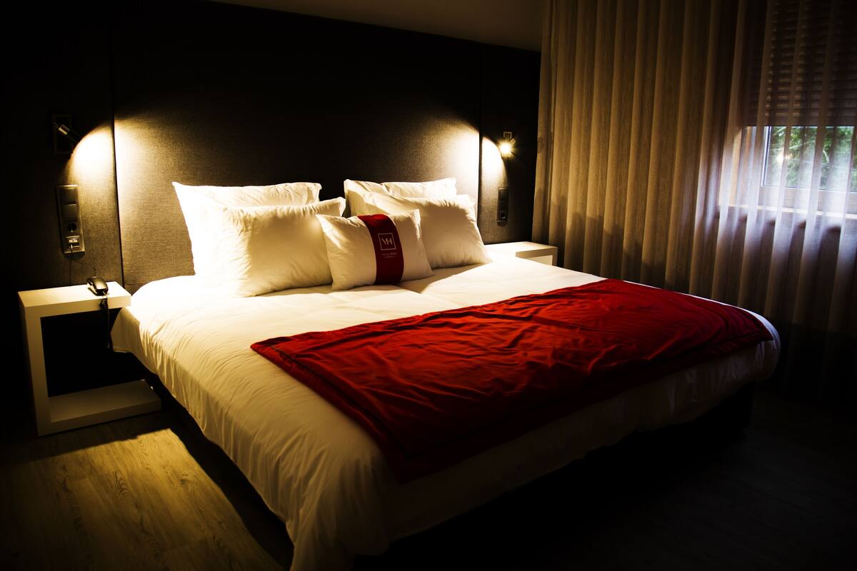 Une chambre d’hôtel avec un grand lit, un plaid rouge, du linge de lit blanc et une table de chevet avec un téléphone et une lampe.