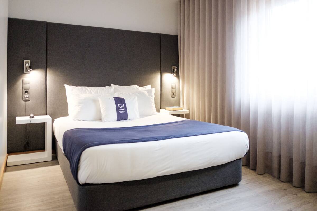 Une chambre d’hôtel moderne avec un grand lit, une tête de lit, des tables de chevet et une fenêtre avec des rideaux.