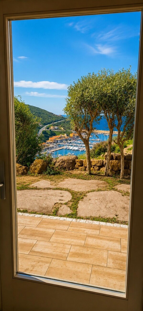 A view through a door of a coastal area with a paved patio, olive trees, and a marina in the background.