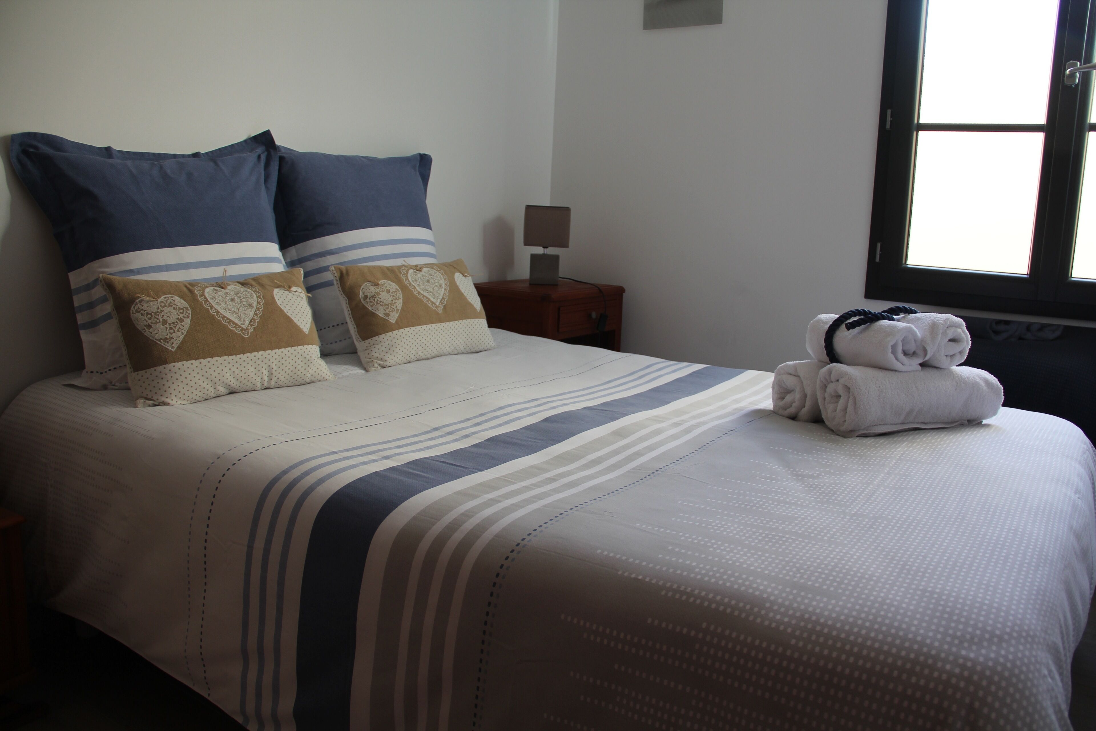A neatly made bed with striped bedding and heart-patterned pillows, a bedside lamp, and a window with natural light.
