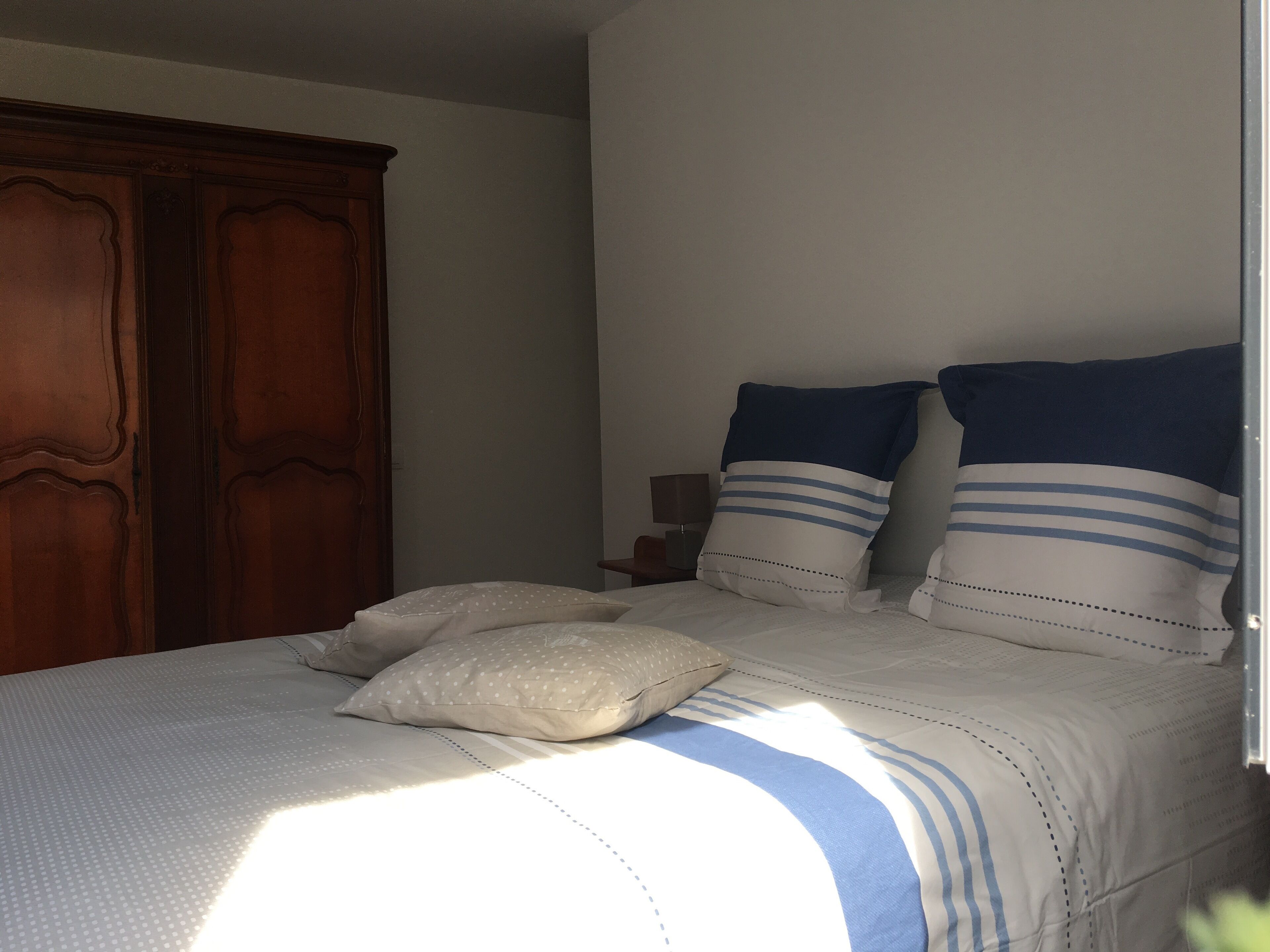A bedroom with a bed, pillows, a wooden wardrobe, and a nightstand with a lamp.