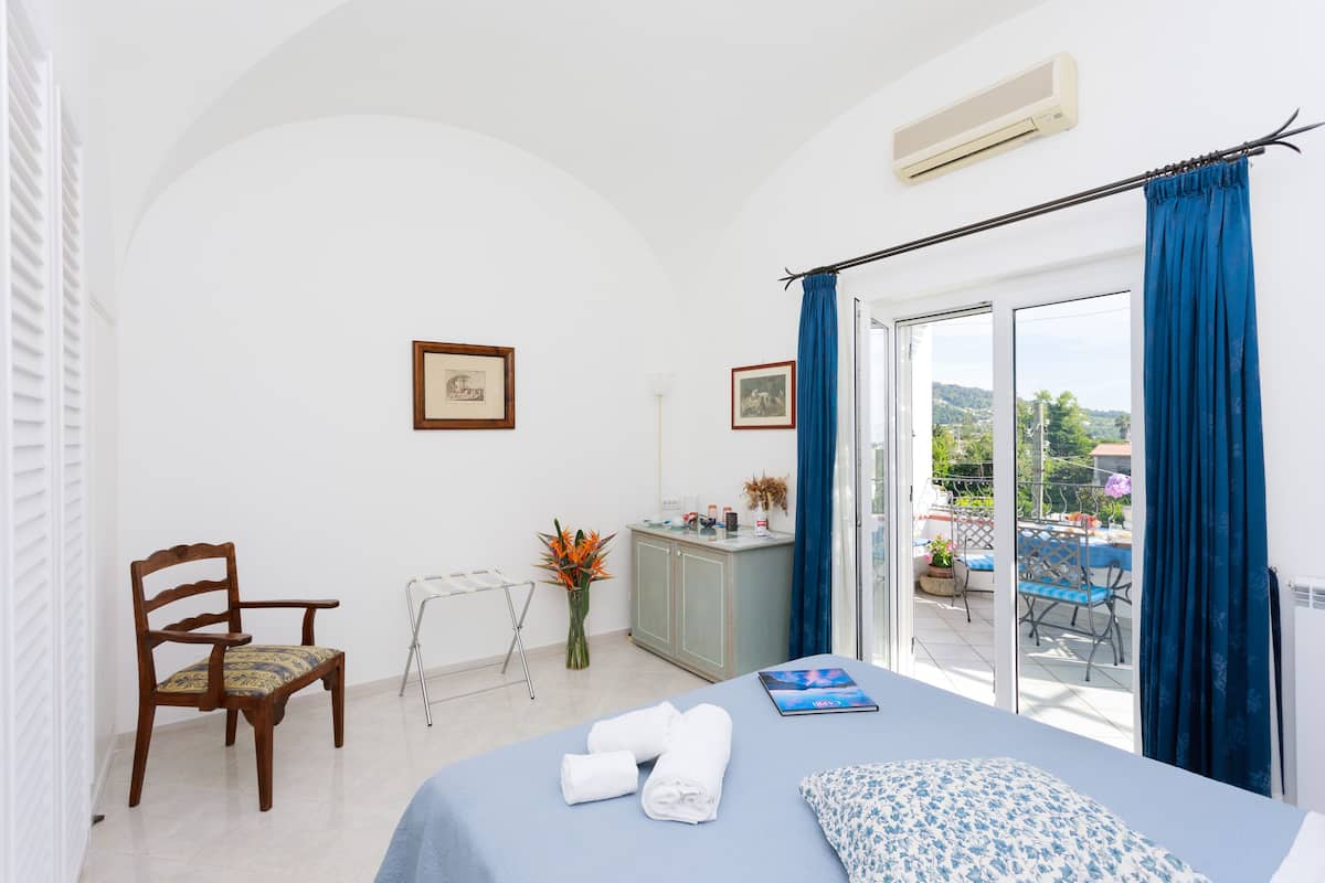 A hotel room with a bed, a chair, a small cabinet, a window with curtains, and a view of the outdoors.